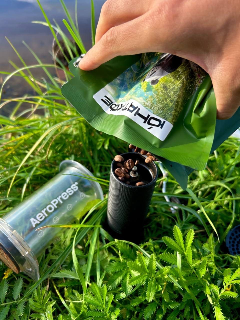Пачка Юнат Кофе, ручная кофемолка и AeroPress среди травы